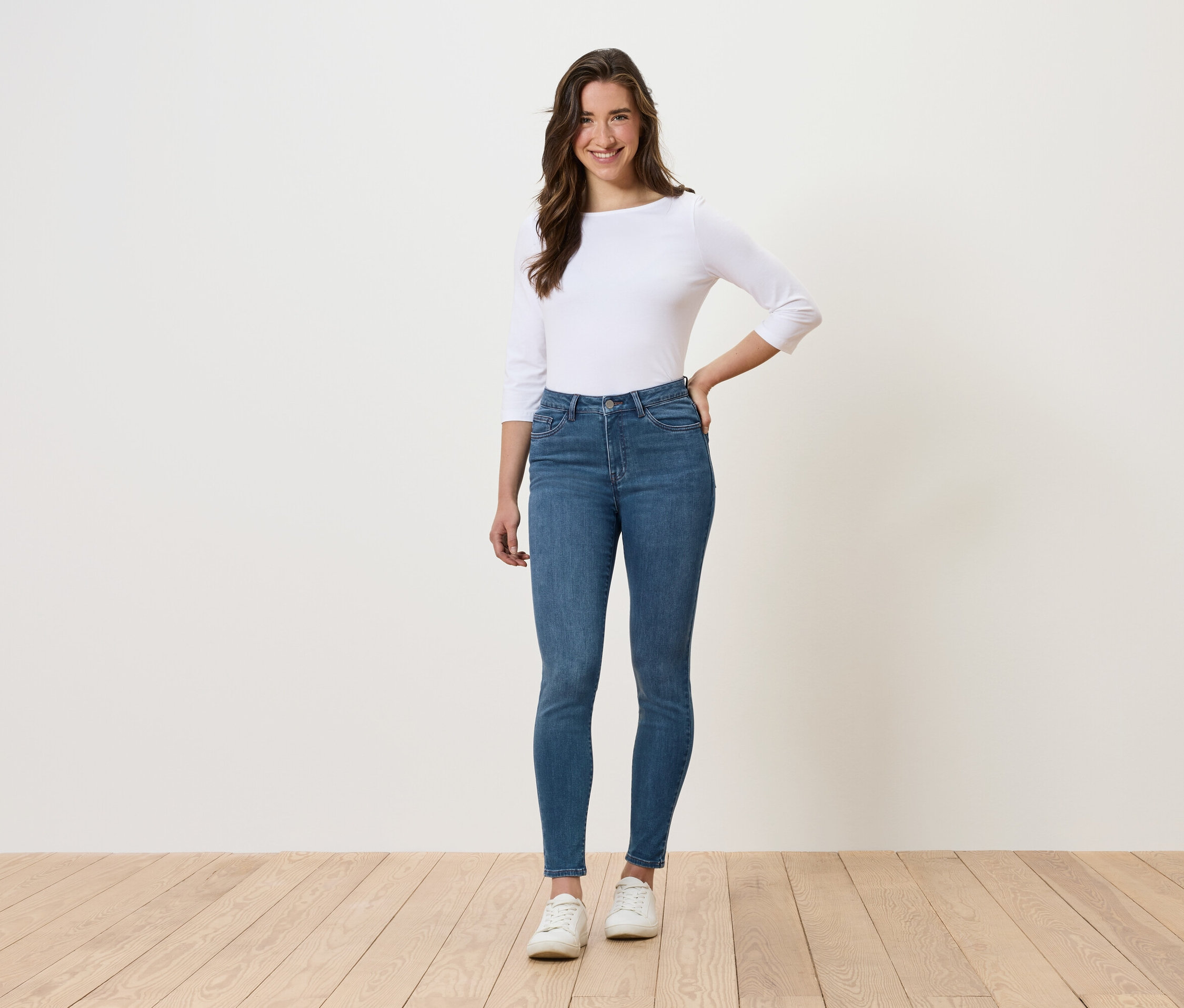 Žena stojí a pózuje v modrých džínách, bílém topu a bílých botách.