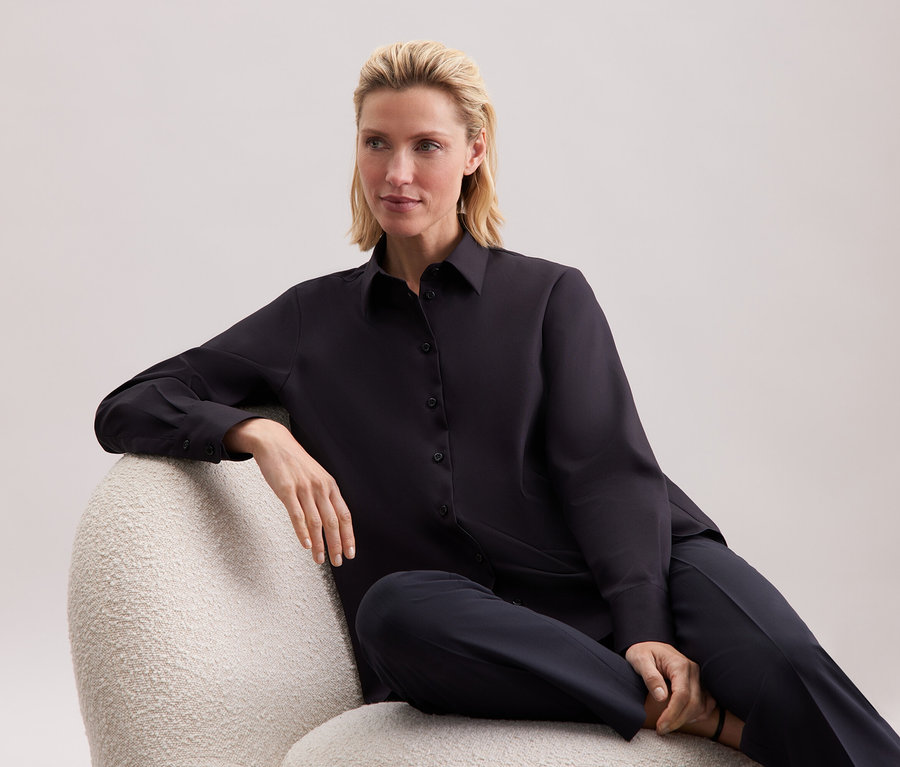 Žena sedí v křesle oblečená v černé košili a kalhotách.