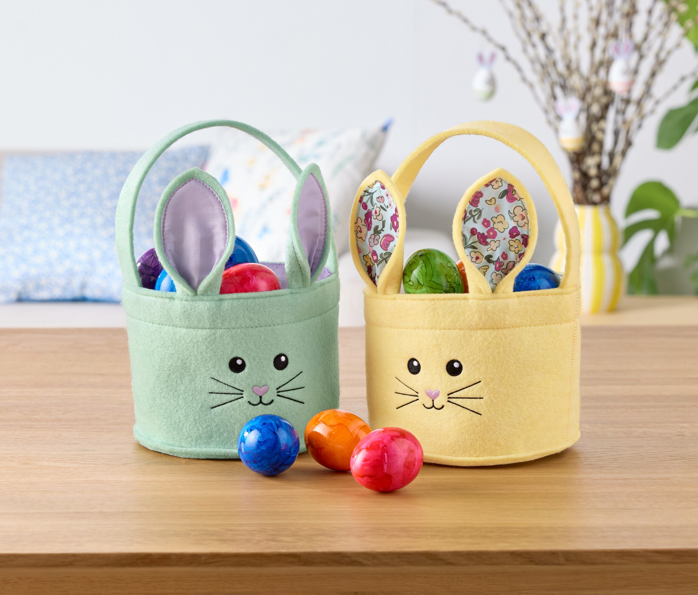 Dva velikonoční košíky ve tvaru zajíčka, jeden mátově zelený a jeden žlutý, plné barevných vajec, stojí na stole.