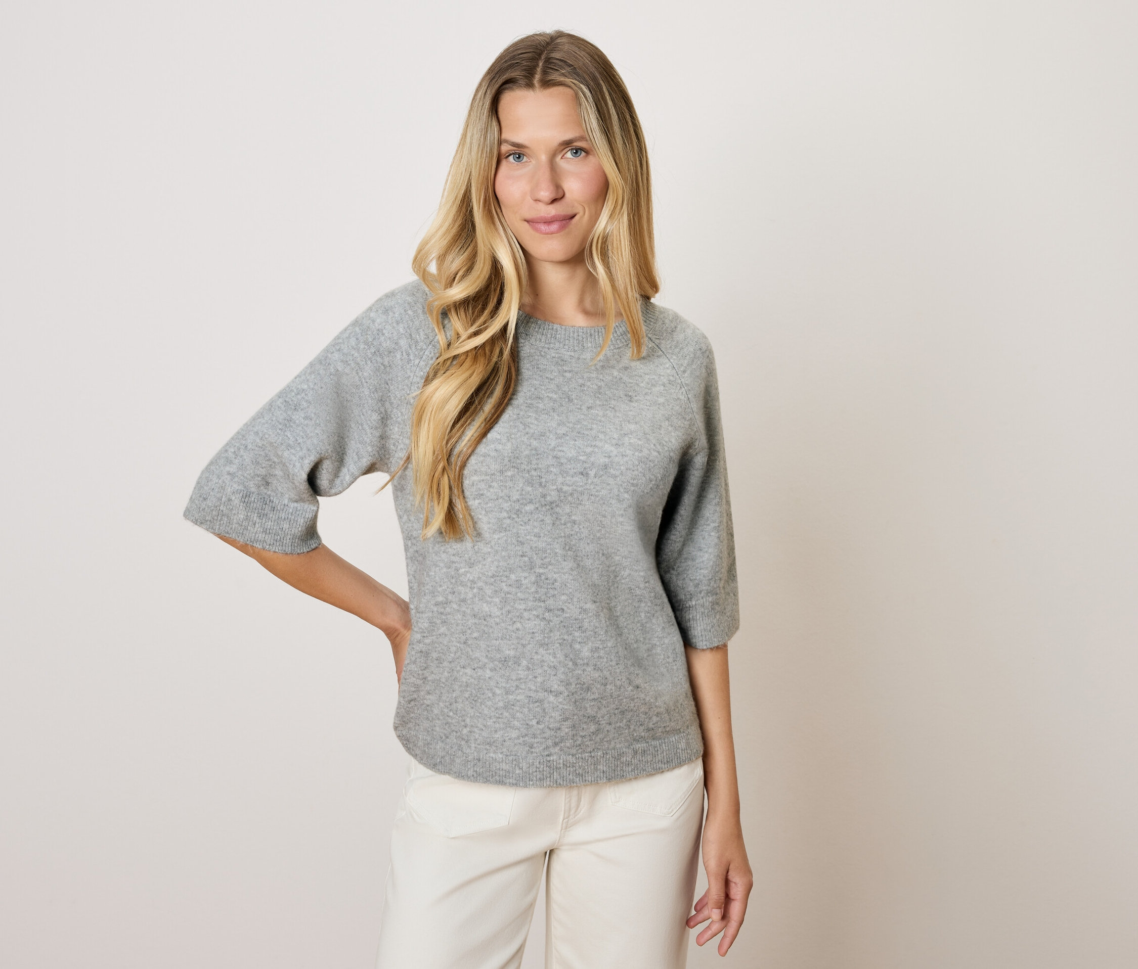 Blondýna v šedém svetru s tříčtvrtečními rukávy a bílých kalhotách pózuje na světlém pozadí.