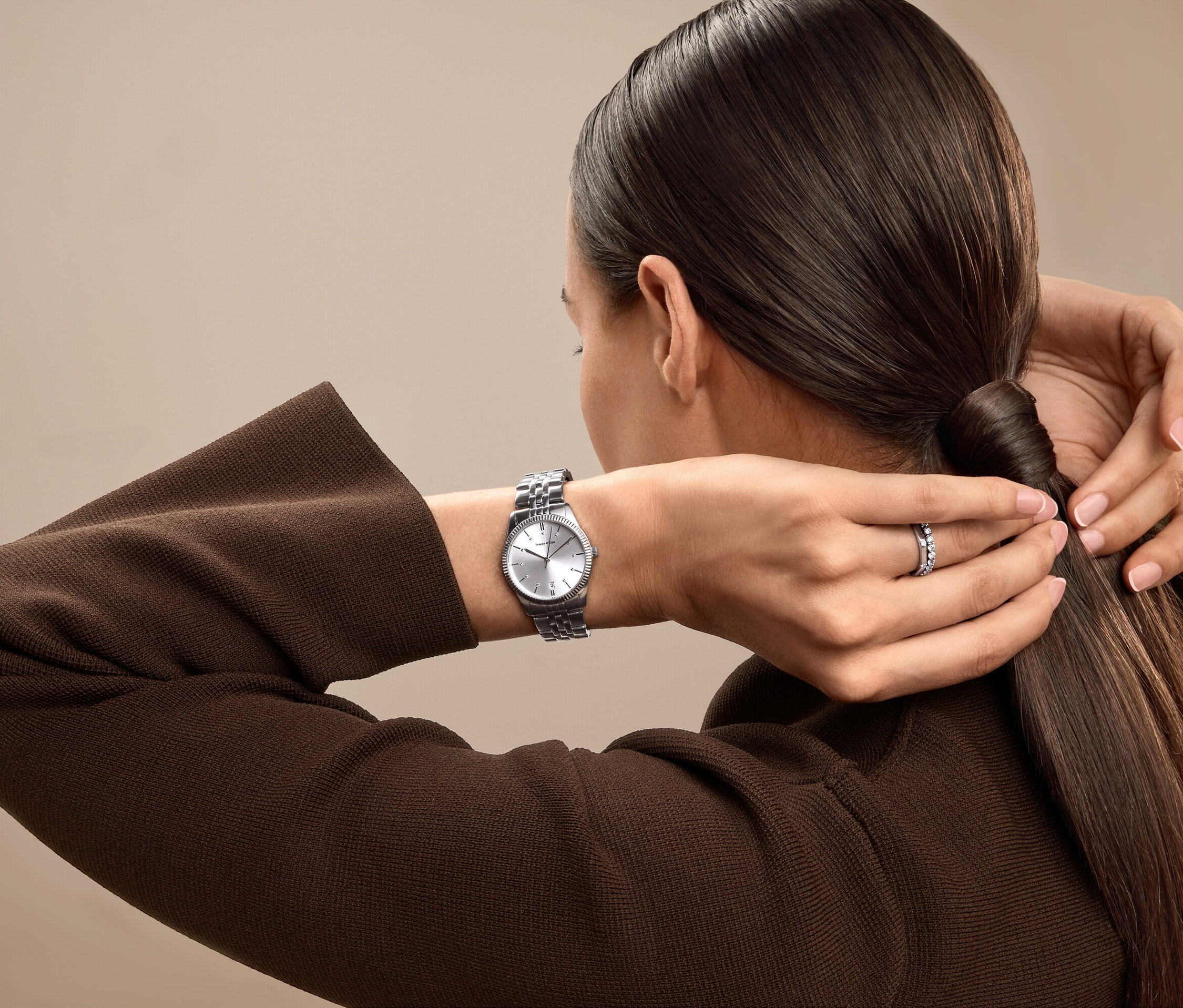 Žena si upravuje vlasy do culíku, na zápěstí má stříbrné hodinky a na prsteníčku prsten.