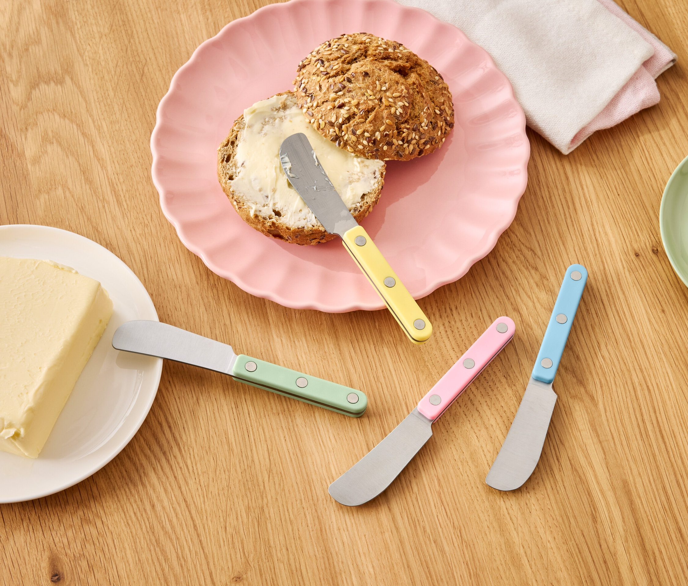 Rohlík s máslem na růžovém talíři vedle kostky másla a barevných nožů na dřevěném stole.