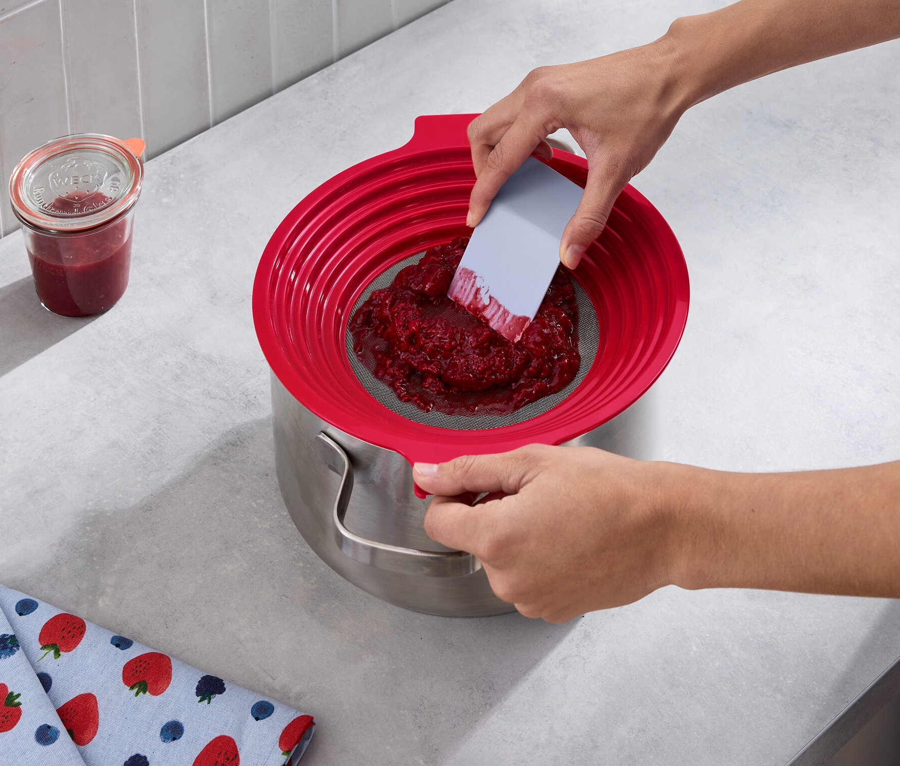 Osoba pasíruje červené ovoce přes 2 v 1: síto na mouku a pasírování do hrnce. Vedle stojí sklenice džemu a leží utěrka s potiskem jahod.