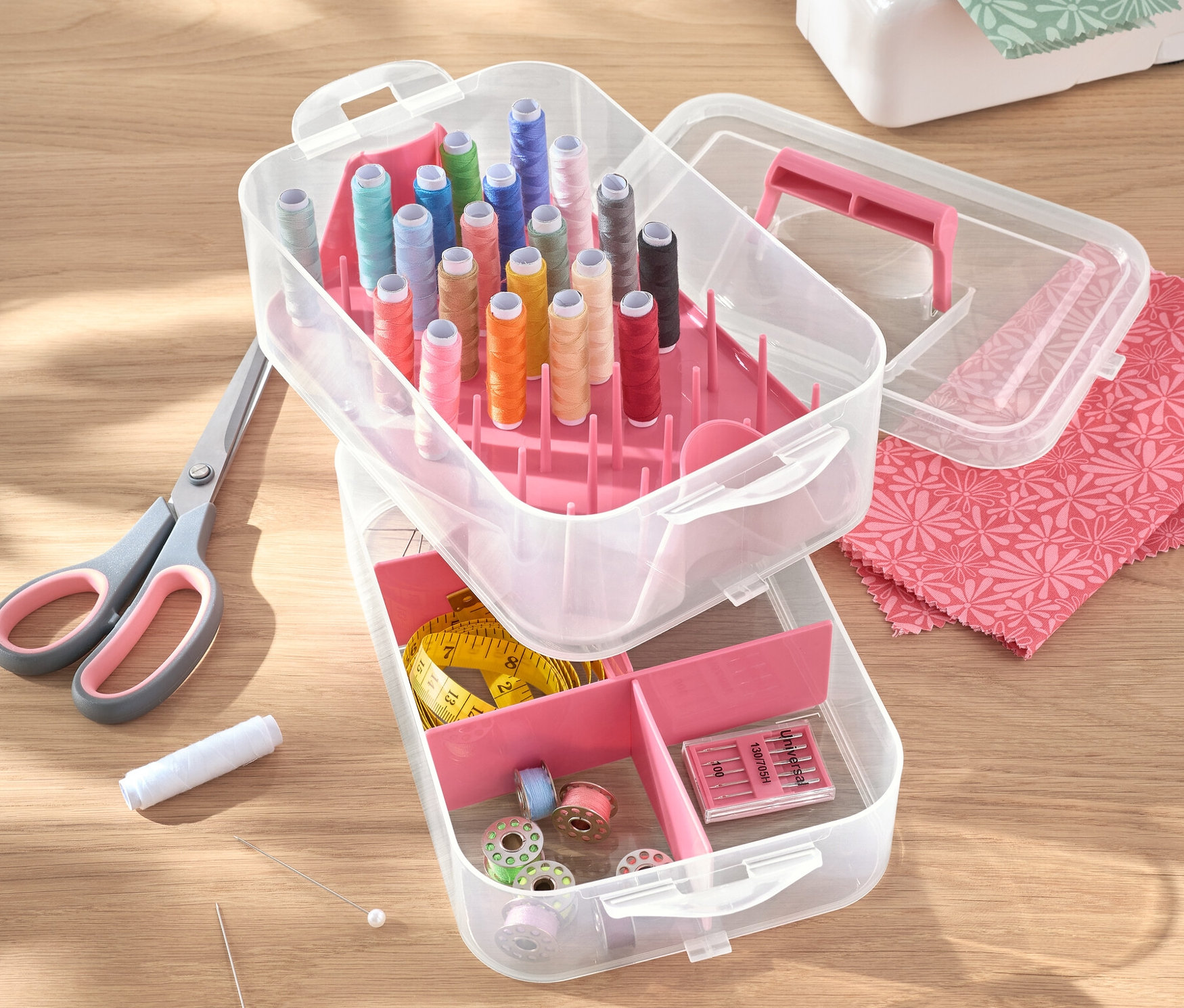 Otevřený plastový box na nářadí s nitěmi, nůžkami, jehlami a šicím strojem na stole.