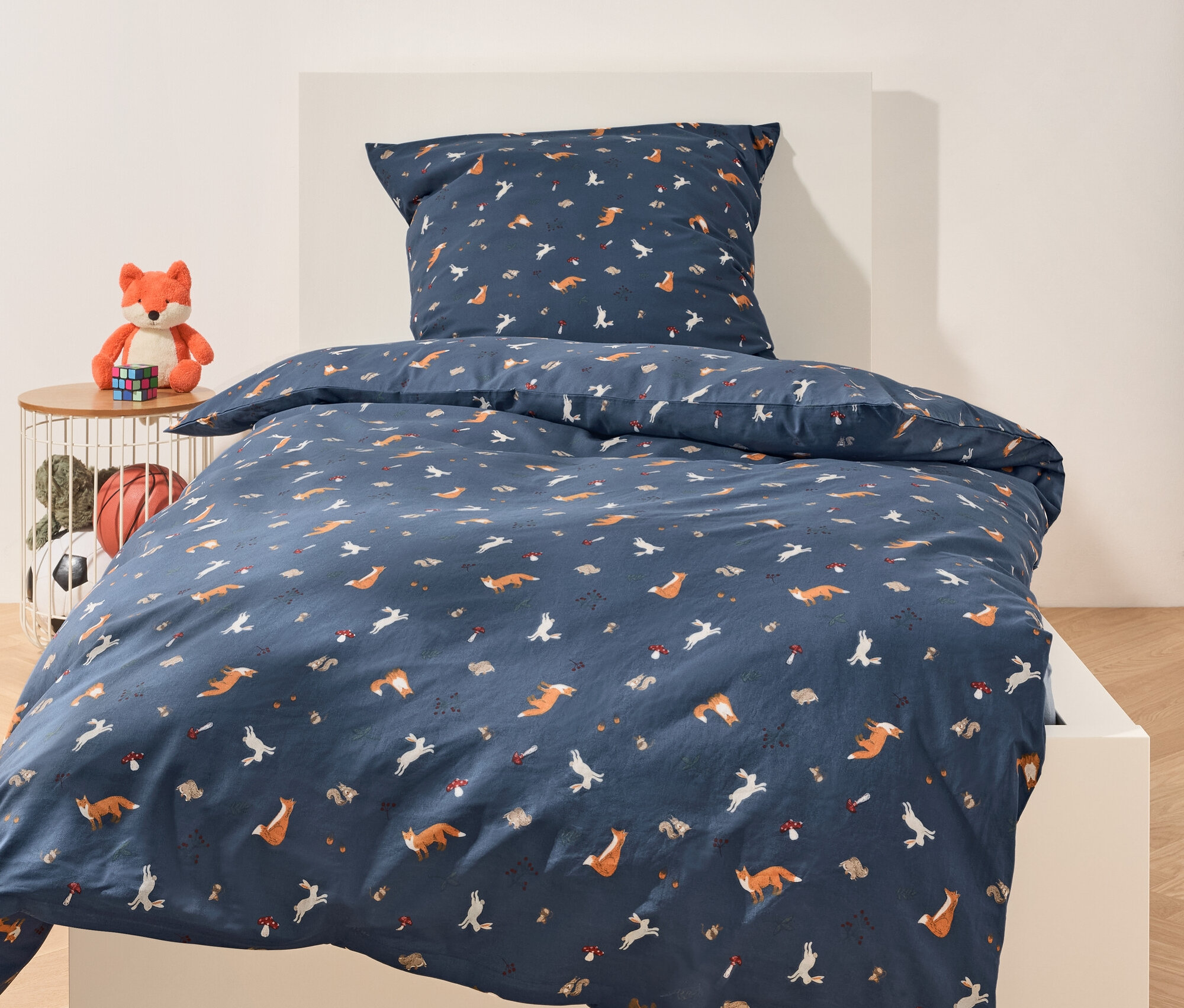 Na bílé posteli je modré povlečení s motivem lišek, králíků, ježků a hub. Vedle postele je stolek s plyšovou liškou a hračkami.