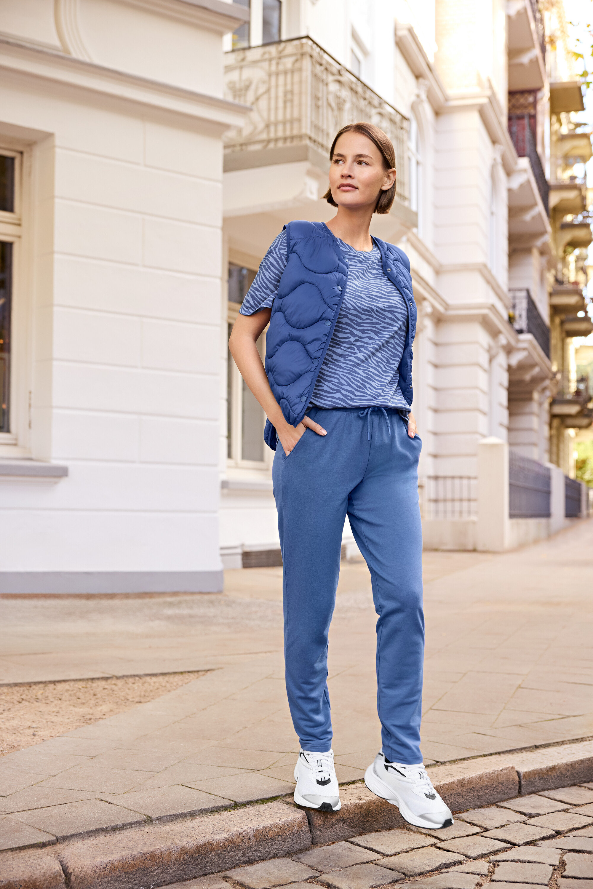 Žena stojí na chodníku a má na sobě modrou vestu, modré tričko se zebřím vzorem a modré tepláky.