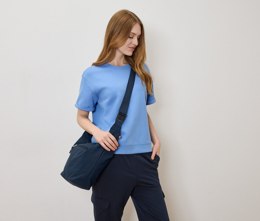 Žena se světle hnědými vlasy, světle modrým tričkem a tmavě modrou taškou přes rameno na krémovém pozadí.