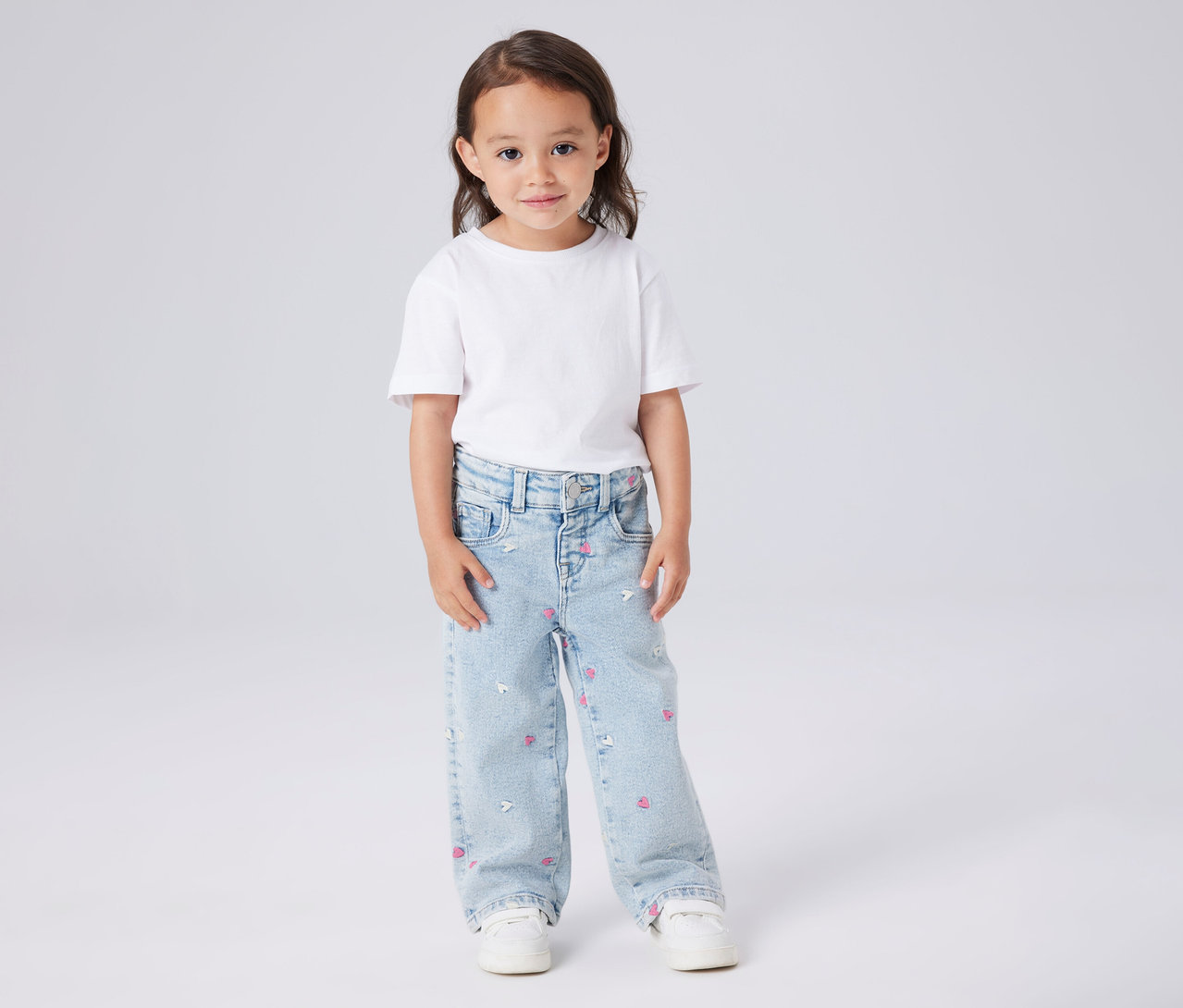 Dívka má na sobě bílé tričko a dětské džíny Wide Leg NAME IT s růžovými srdíčky a bílými myškami.