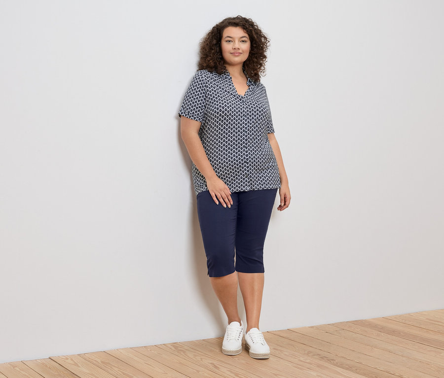 Žena s kudrnatými vlasy se opírá o zeď a má na sobě modro-bílou vzorovanou blůzu, modré capri kalhoty a bílé tenisky.