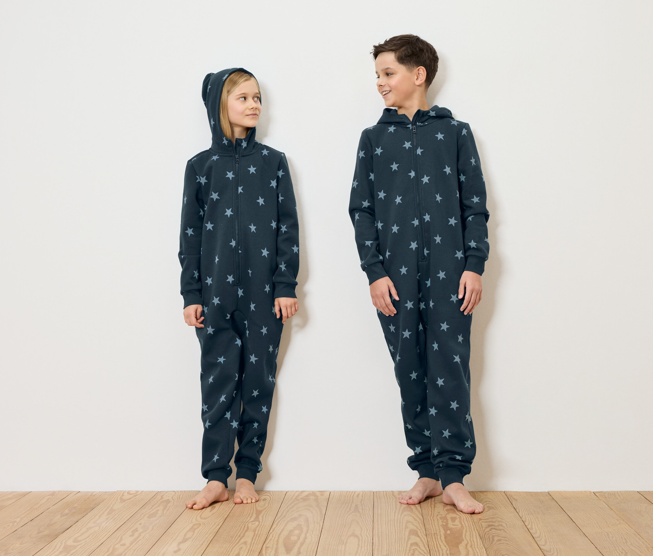 Dívka a chlapec stojí bosí před bílou stěnou a mají na sobě tmavě modré pyžama s hvězdami.
