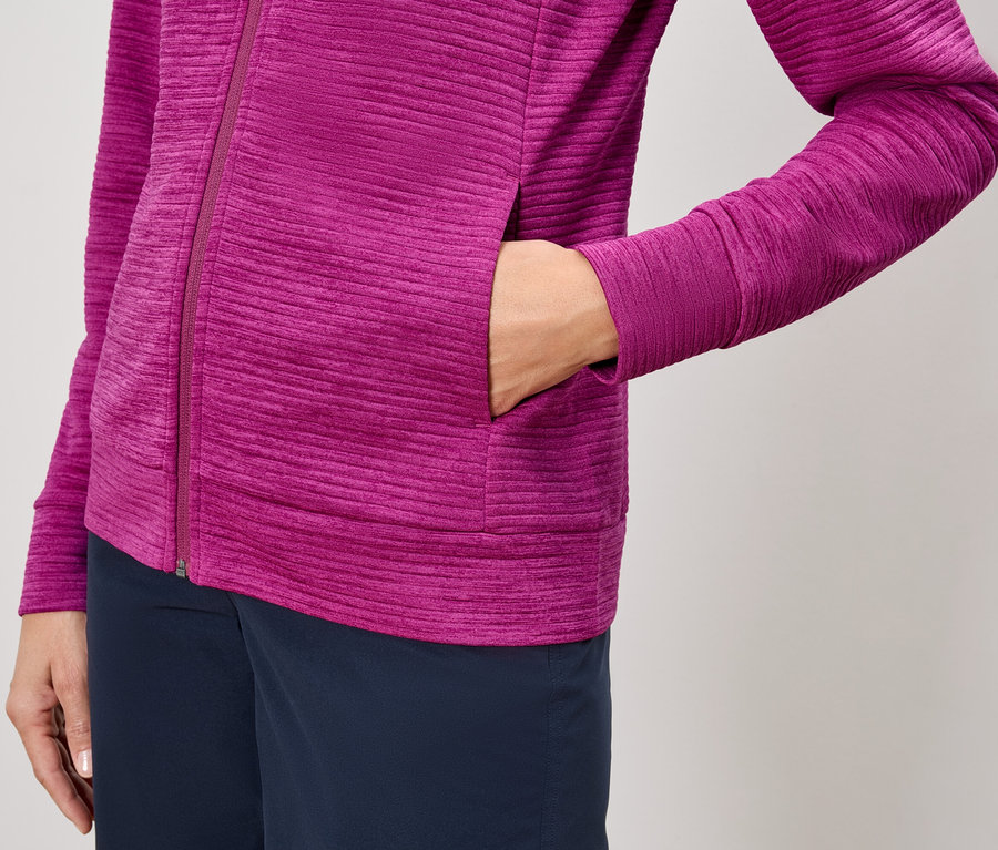 Detail osoby v růžové bundě na zip a tmavě modrých kalhotách, s rukou v kapse bundy.