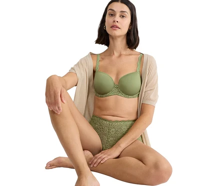 Žena sedí se zkříženýma nohama a má na sobě zelenou podprsenku a zelené krajkové kalhotky, spolu s béžovým svetrem.
