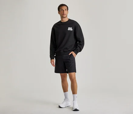 Muž má na sobě černou mikinu s nápisem „RUN CLUB“, černé šortky a bílé tenisky. Jeho levá ruka je v kapse šortek.