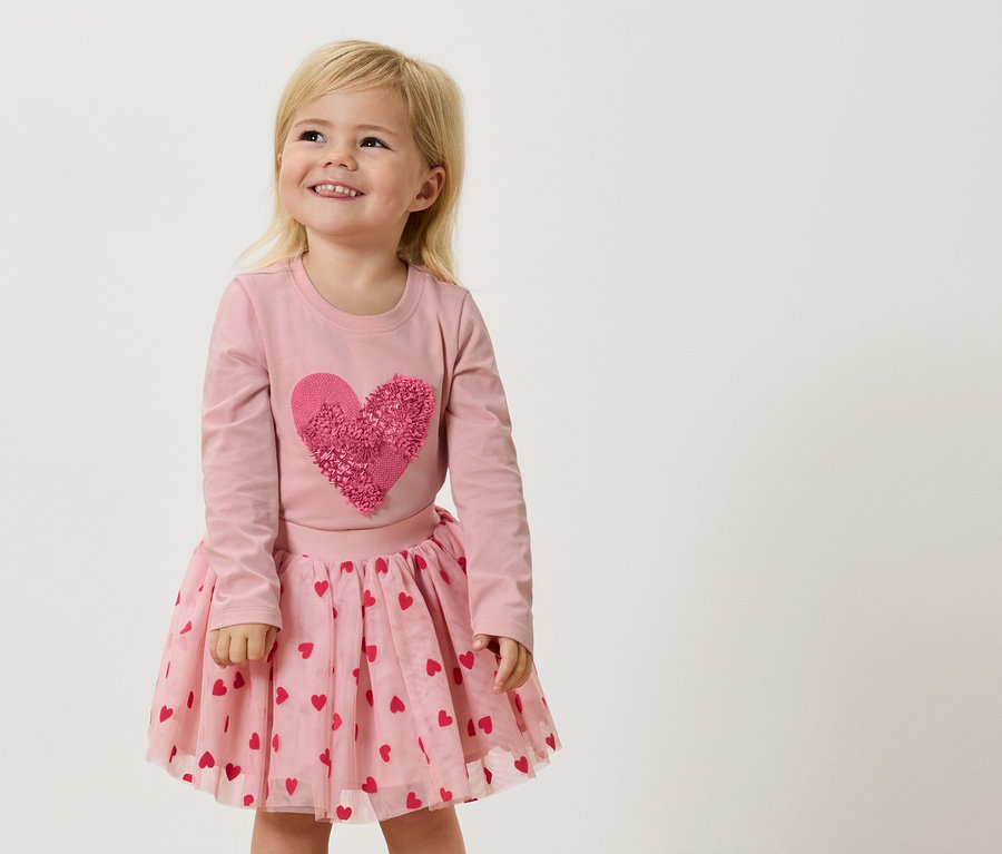 Malá blondýnka má na sobě růžové tričko s dlouhým rukávem s velkým růžovým srdcem a růžovou tylovou sukni s červenými srdíčky.