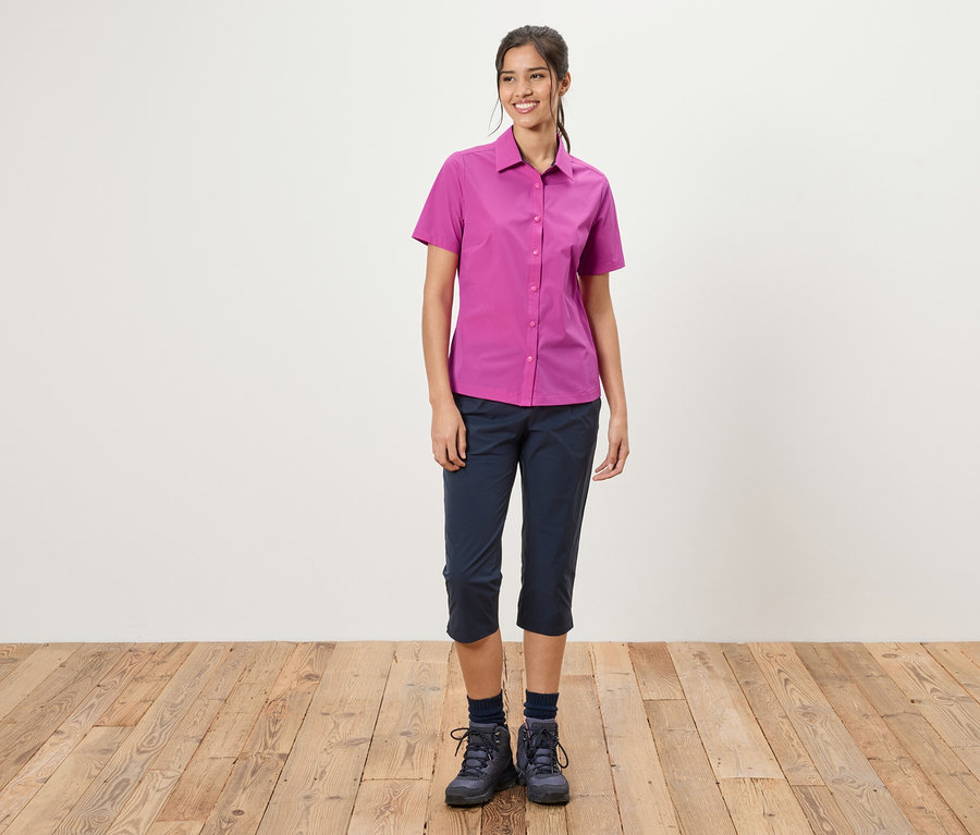 Žena stojí před bílou stěnou. Má na sobě růžovou košili, modré tříčtvrťáky a trekové boty.