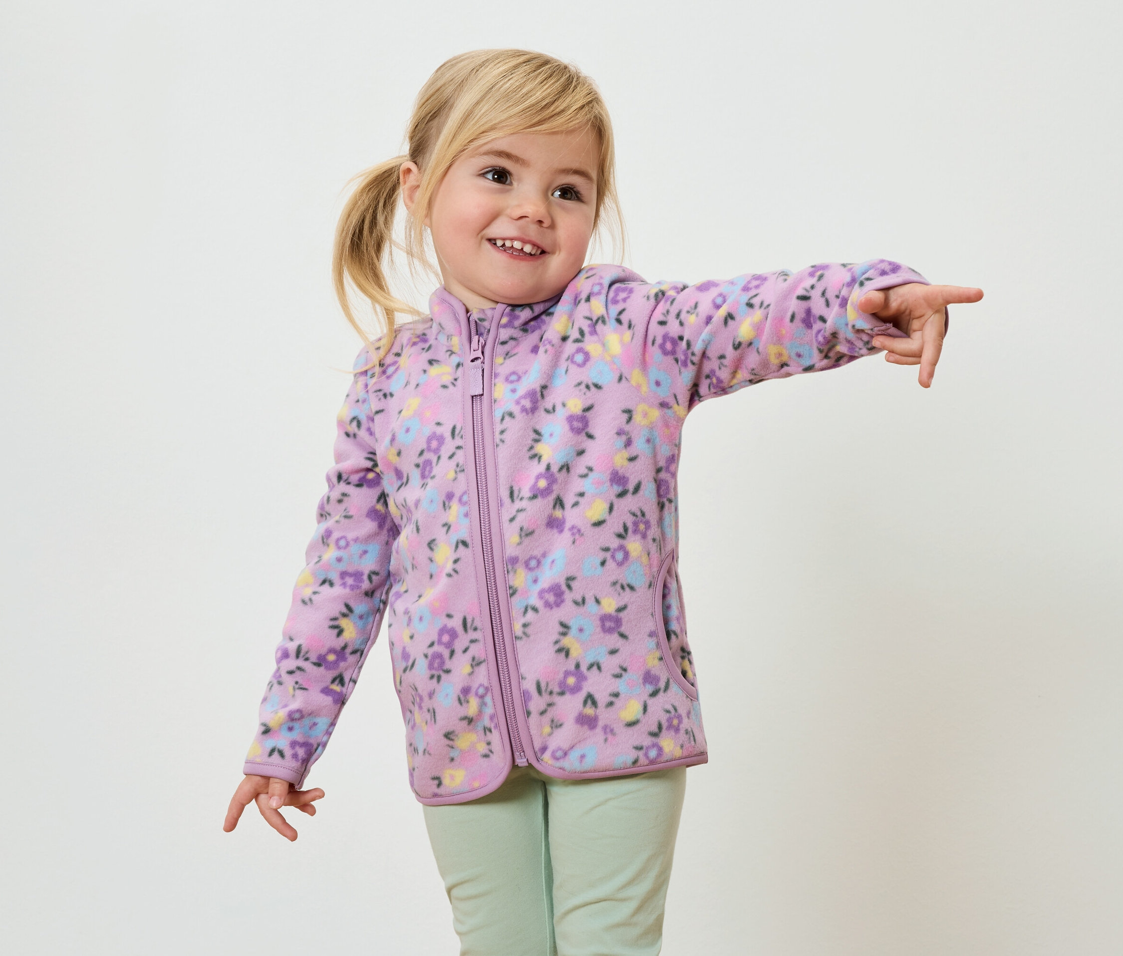 Usmívající se dívka s blond copánky, oblečená do fialové fleecové bundy s květinovým vzorem a světle zelených kalhot, ukazuje prstem.