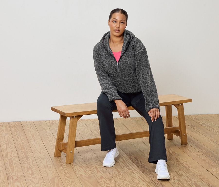 Žena sedí na dřevěné lavičce a má na sobě šedou fleecovou mikinu s leopardím vzorem, růžový top, tmavé kalhoty a bílé tenisky.