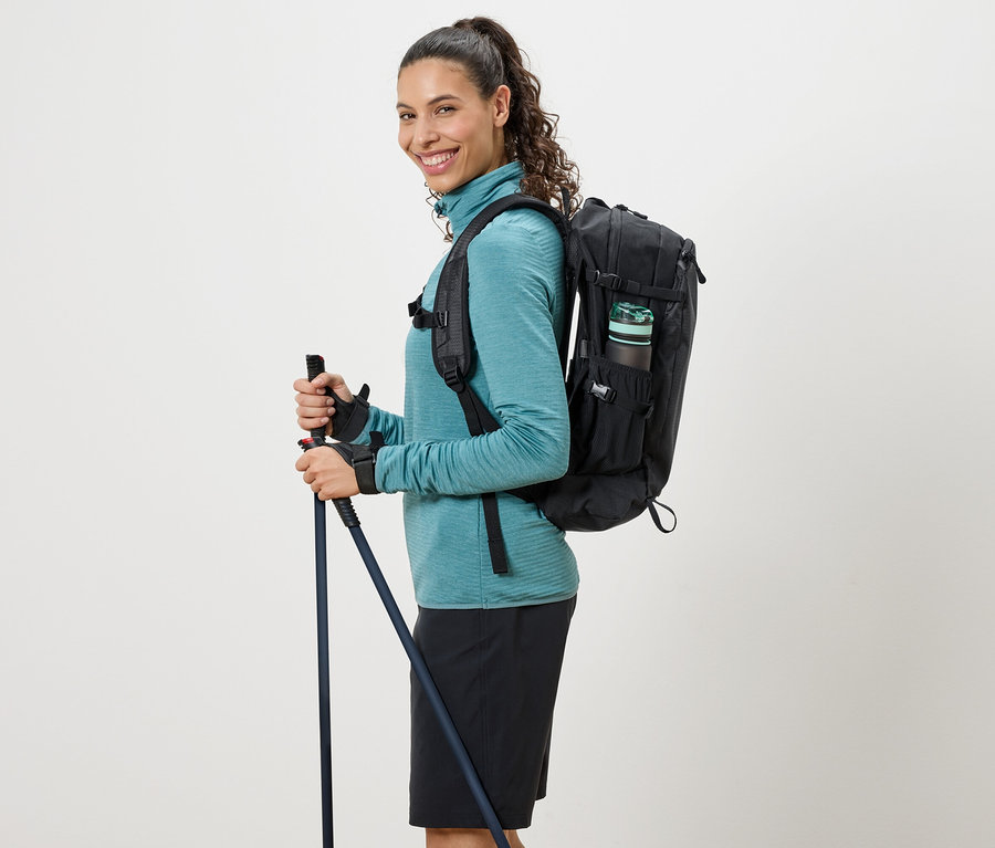 Žena v tyrkysovém roláku, s černým batohem a trekovými holemi.