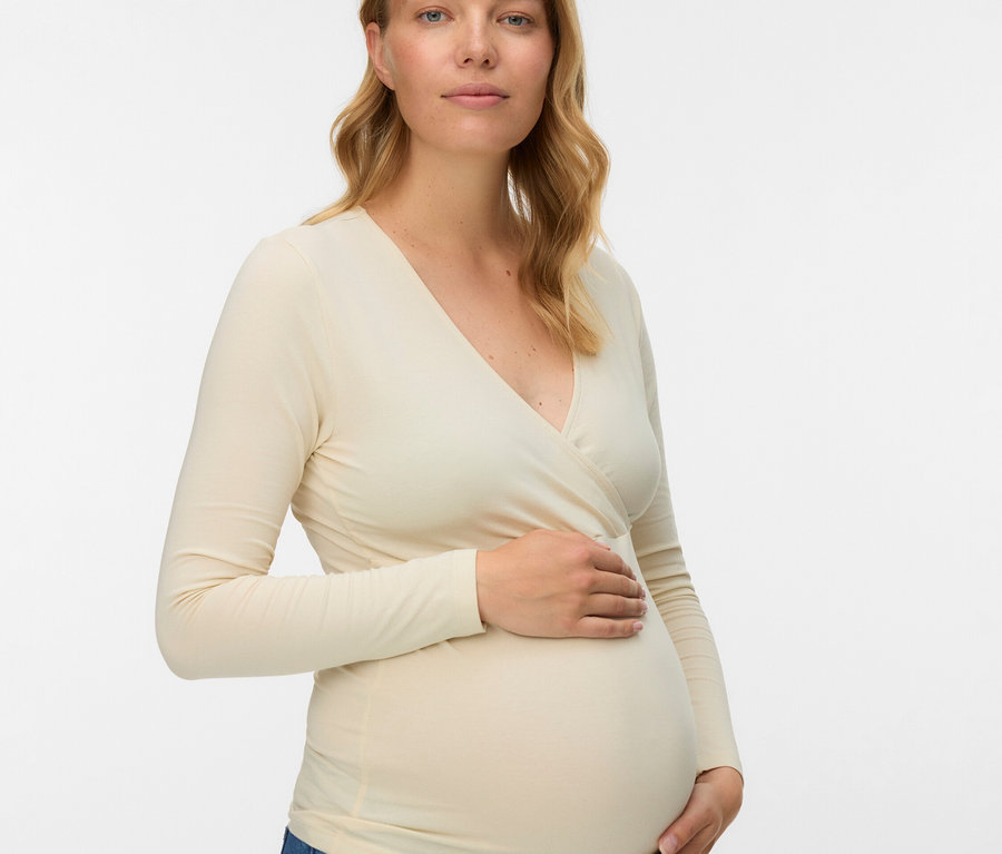 Těhotná žena s blond vlnitými vlasy má na sobě blůzu s dlouhým rukávem a džíny.