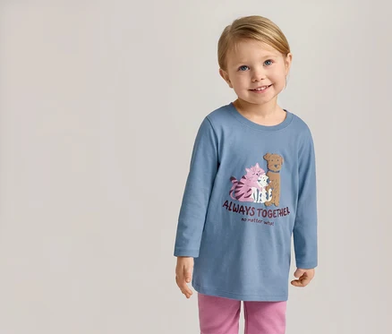Usmívající se dívka v modrém tričku s dlouhým rukávem s motivem koček a psů a nápisem „Always Together“ a růžových kalhotách.