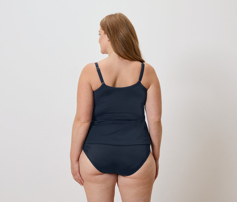 Pohled zezadu na ženu s dlouhými hnědými vlasy v námořnicky modrém tankini a plavkách na bílém pozadí.