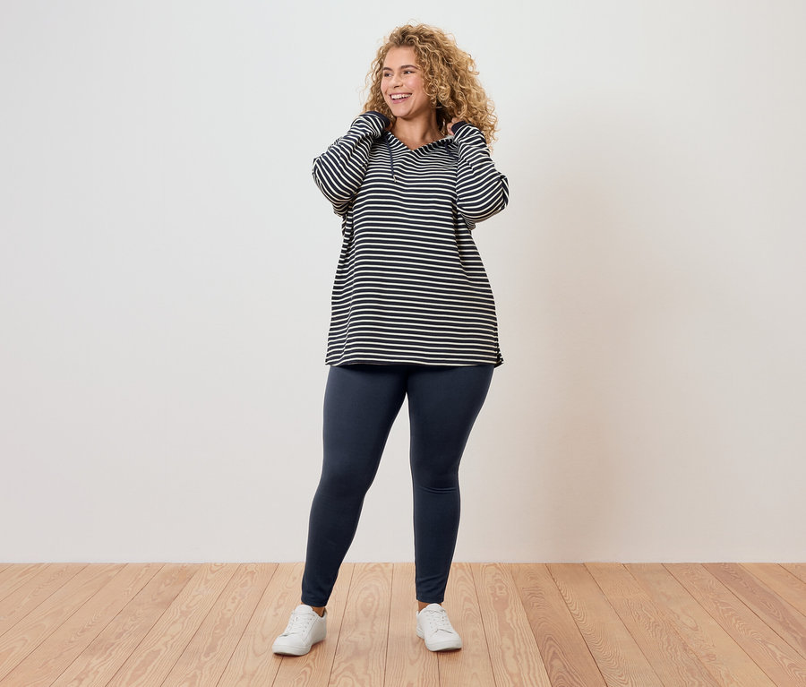 Žena s kudrnatými vlasy v pruhovaném topu, legínách a bílých teniskách.