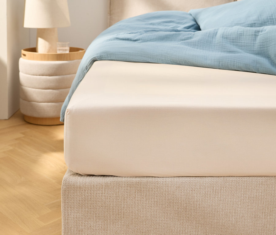Detailní záběr béžového prostěradla s gumou na posteli se světle modrým ložním prádlem. Vedle stojí noční stolek s lampou.