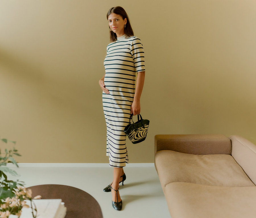 Těhotná žena v pruhovaných šatech MAMALICIOUS »MLLANE« drží kabelku a rukou se dotýká břicha. Má na sobě černé boty.