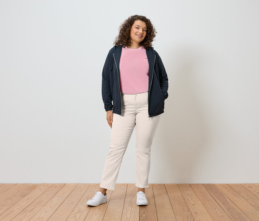 Žena s kudrnatými vlasy stojí před zdí a má na sobě tmavě modrou mikinu s kapucí, růžové tričko, krémové kalhoty a bílé tenisky.