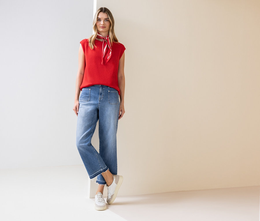 Žena stojí a má na sobě červený top, šátek, džíny a tenisky.