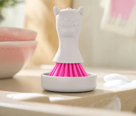 Bílý kartáč s růžovými štětinami ve tvaru lamy stojí v bílé misce na dřezu se zbytky mýdla.