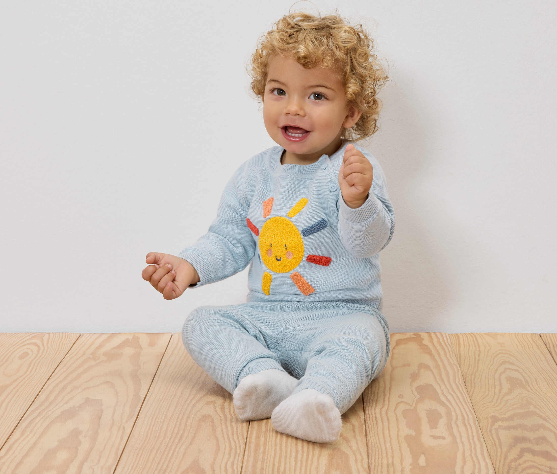 Usmívající se dítě s blond kudrlinami sedí na dřevěné podlaze a má na sobě světle modrý svetr s vyšitým sluncem a odpovídající kalhoty.