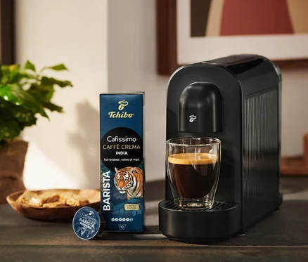 Černý kávovar Tchibo Cafissimo se sklenicí kávy, vedle balení kávových kapslí Tchibo Cafissimo Caffè Crema India a jedna kapsle.