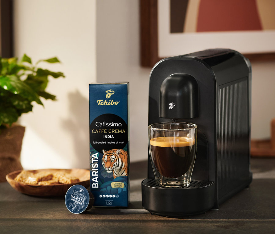 Černý kávovar Tchibo Cafissimo se sklenicí kávy, vedle balení kávových kapslí Tchibo Cafissimo Caffè Crema India a jedna kapsle.