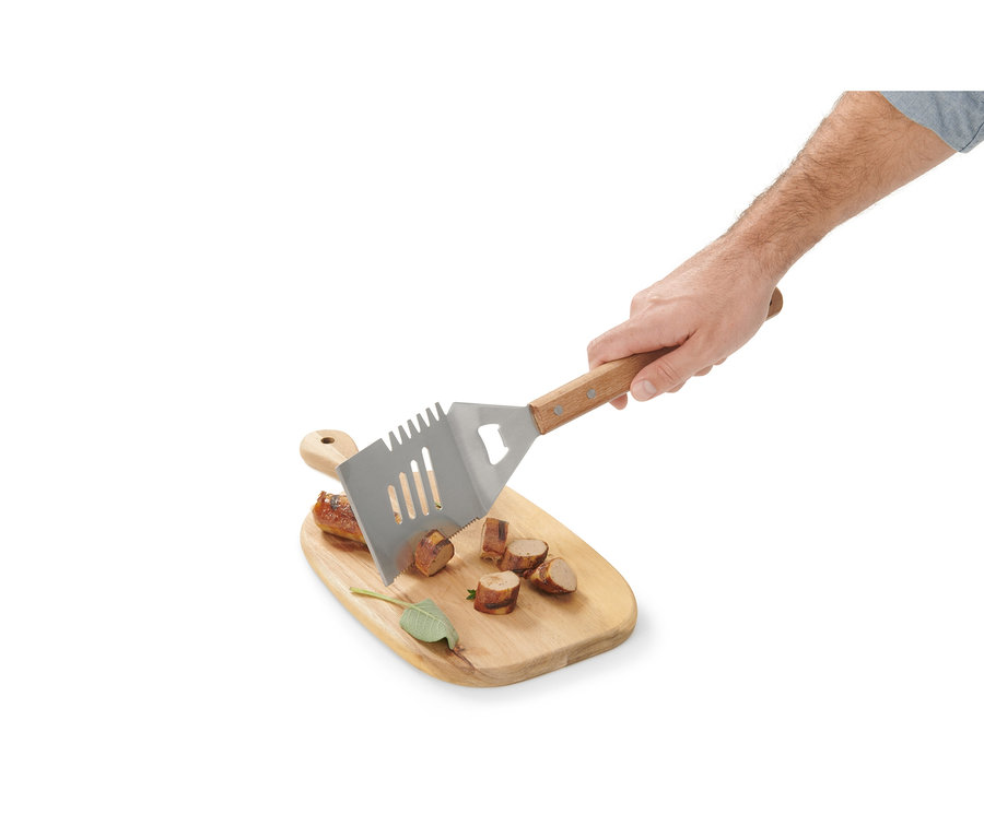Ruka drží multifunkční BBQ náčiní nad dřevěným prkénkem s nakrájenou klobásou.