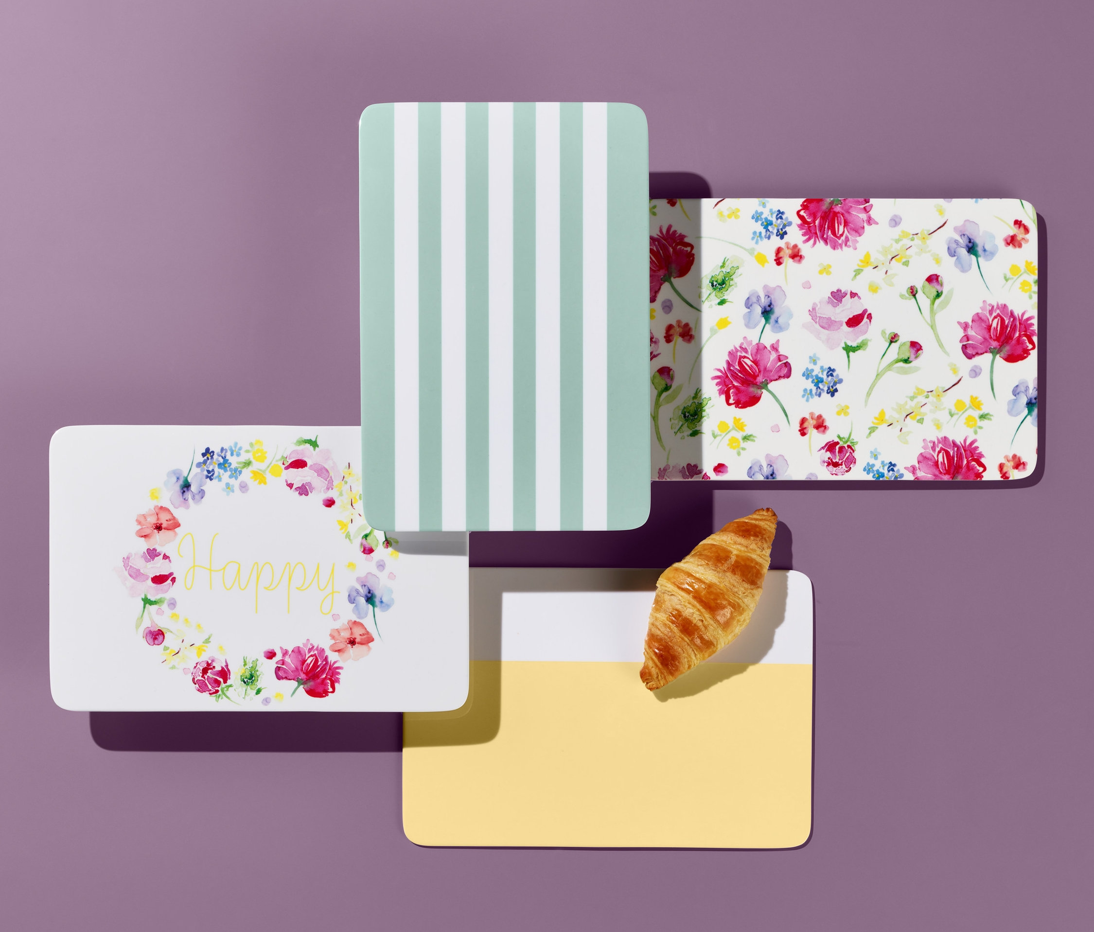 Čtyři krájecí prkénka s různými vzory na fialovém pozadí, croissant.