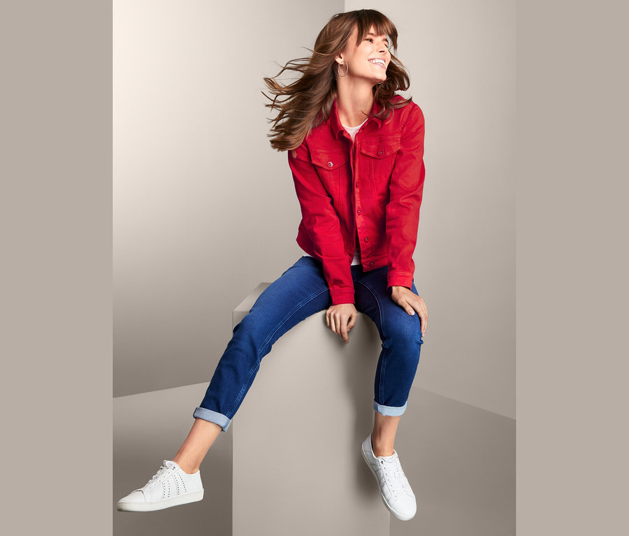 Žena sedí a má na sobě džínovou bundu, džíny a tenisky.