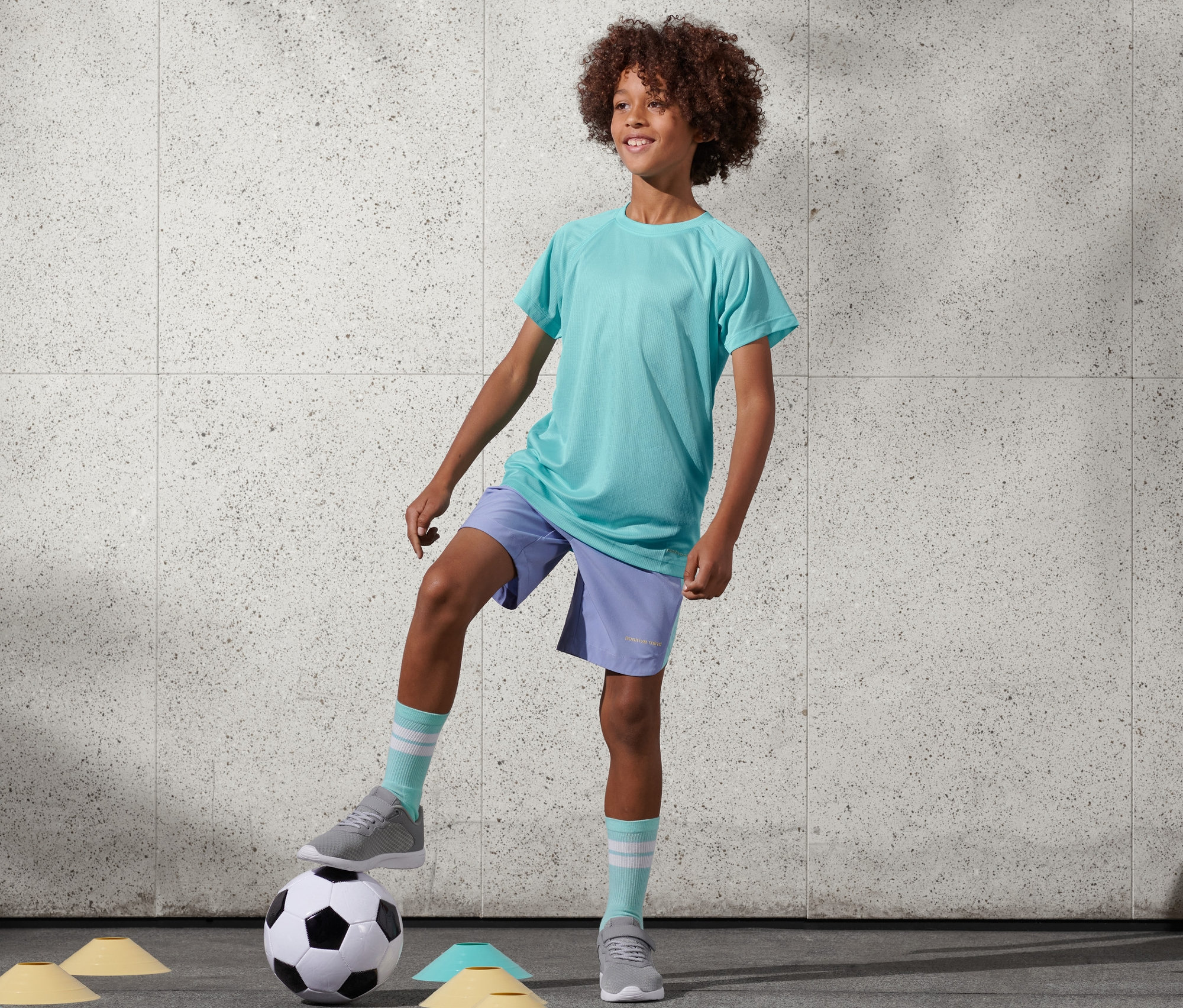 Chlapec s afro účesem stojí s jednou nohou na fotbalovém míči. Má na sobě tyrkysové funkční tričko z recyklovaného materiálu, 2 ks a fialové šortky.