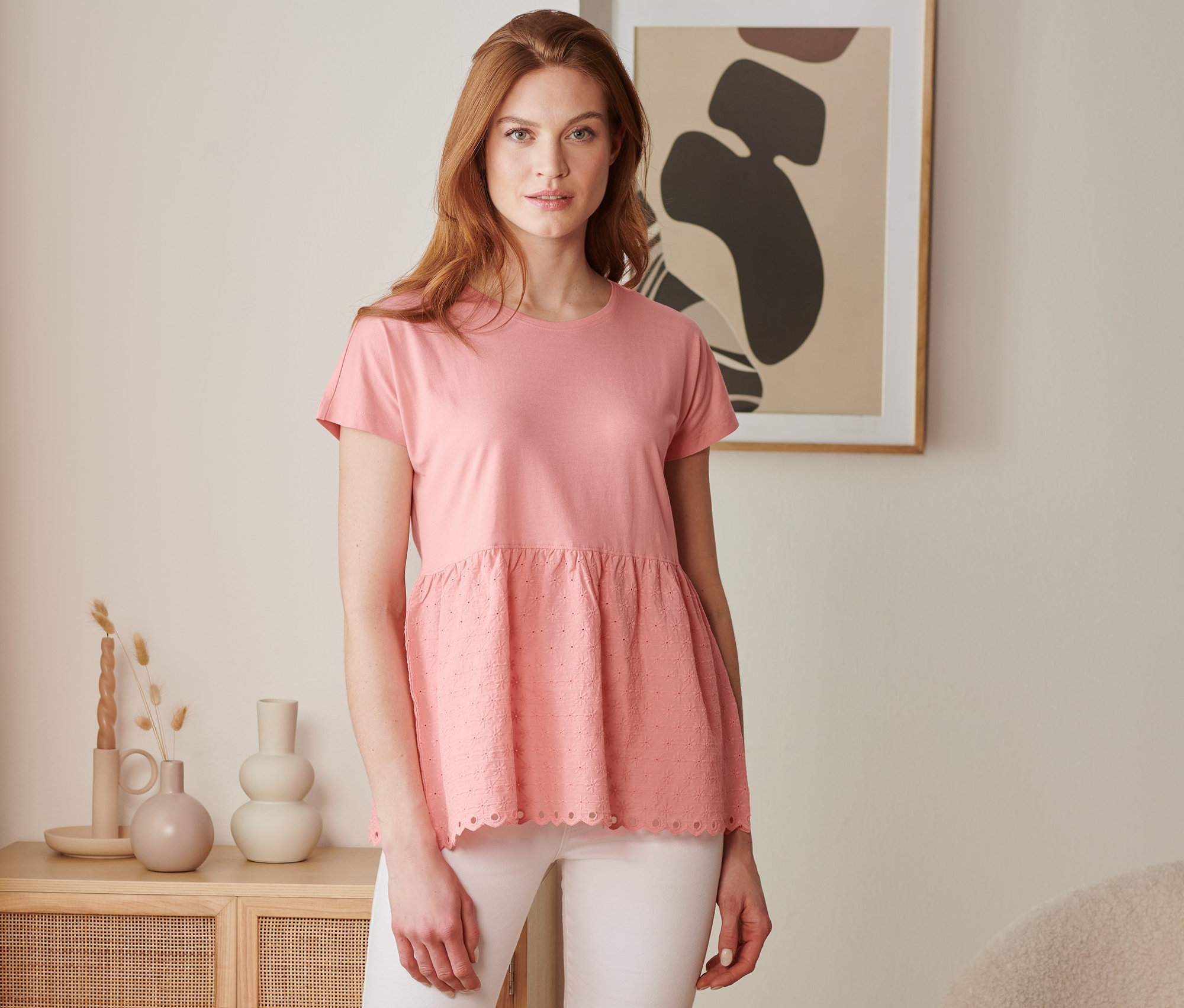 Žena má na sobě korálové tričko z kombinace materiálů s výšivkou a bílé kalhoty.