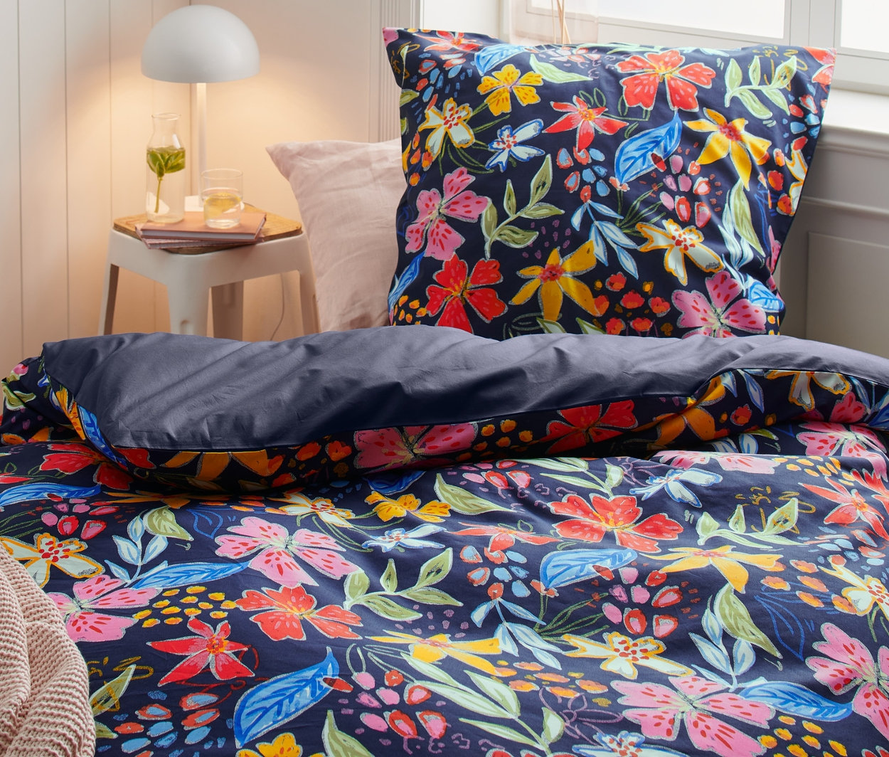 Postel s perkálovým povlečením s květinovým vzorem, včetně velikostí na dvoulůžko, standardní a nadměrné velikosti. Na nočním stolku lampa.