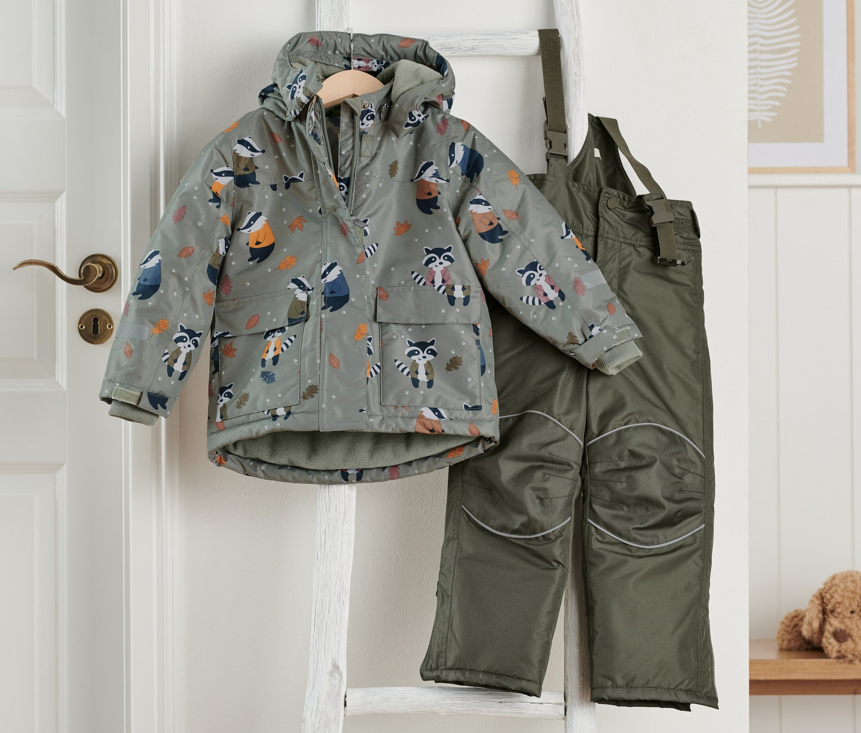 Dvoudílná zimní kombinéza olivově zelené barvy visí na žebříku.