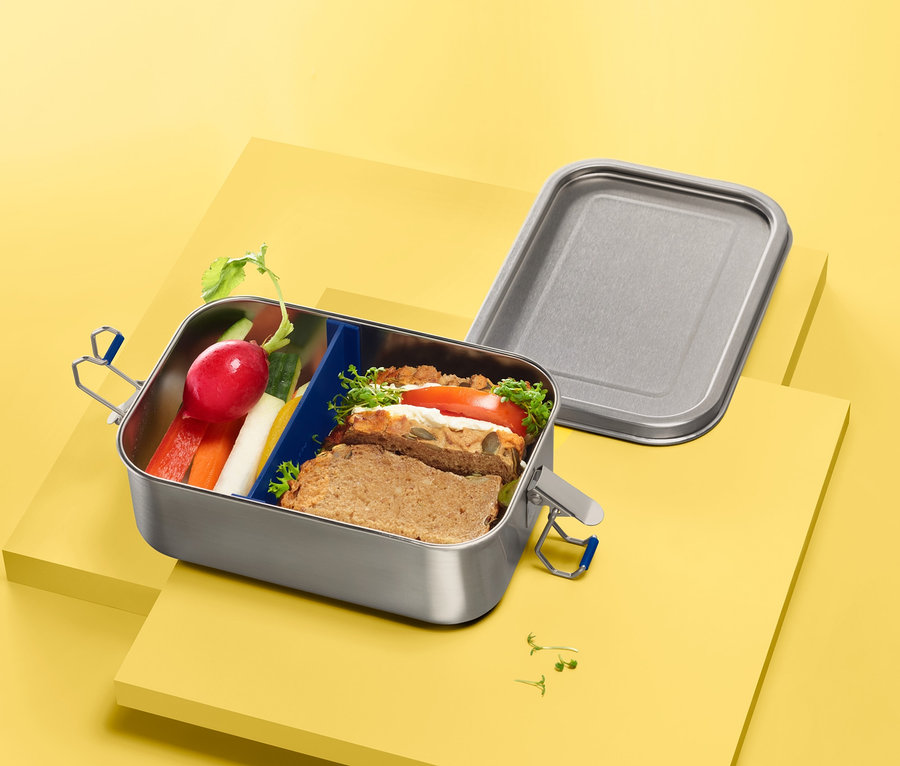 Nerezová cestovní dóza na svačinu s obsahem, položená na žlutém podkladu.