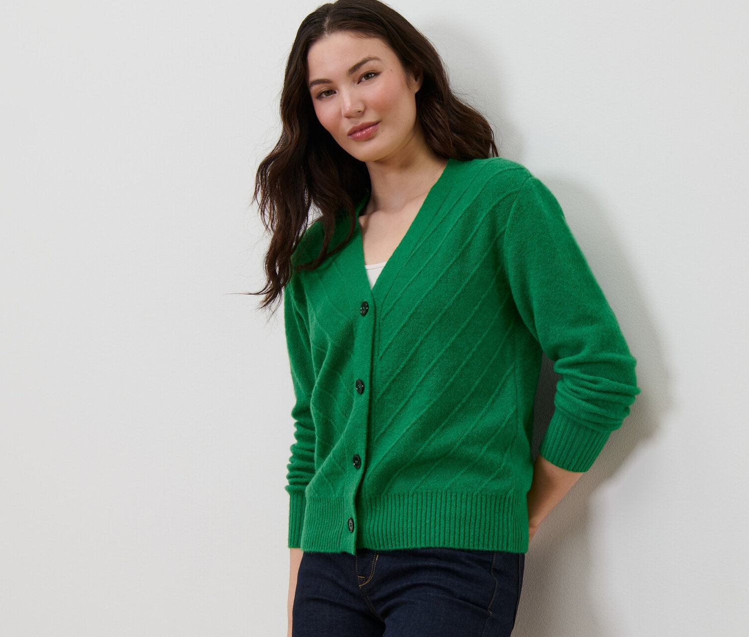 Žena pózuje v zeleném kardiganu s pletenou strukturou a tmavě modrých džínách.