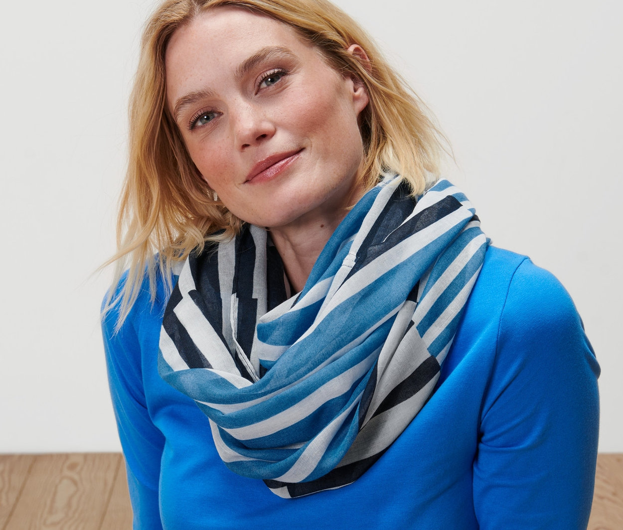 Blondýna sedí na podlaze a má na sobě pruhovanou kruhovou šálu.