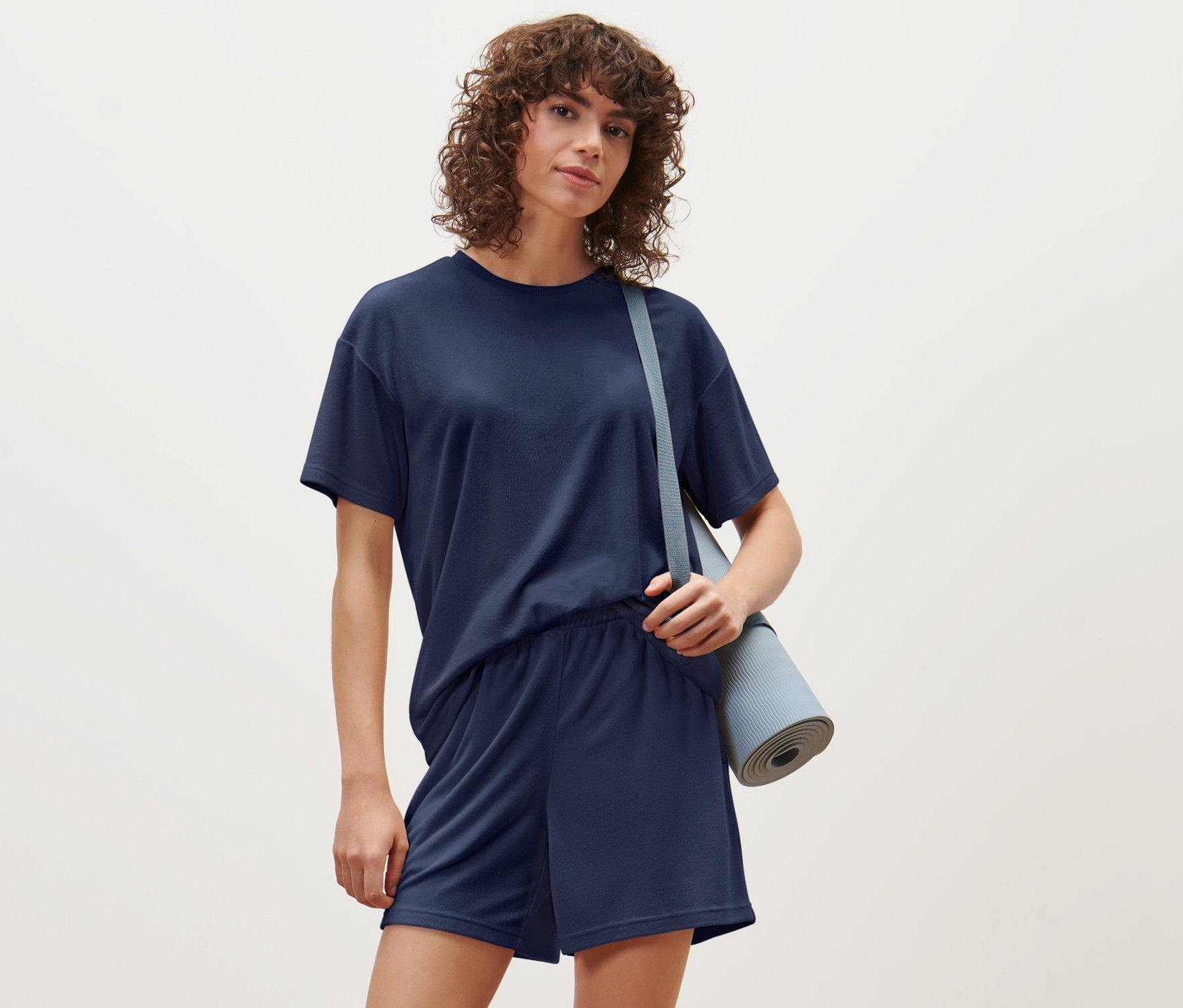 Žena má na sobě sportovní tričko z froté a šortky a přes rameno podložku na cvičení.