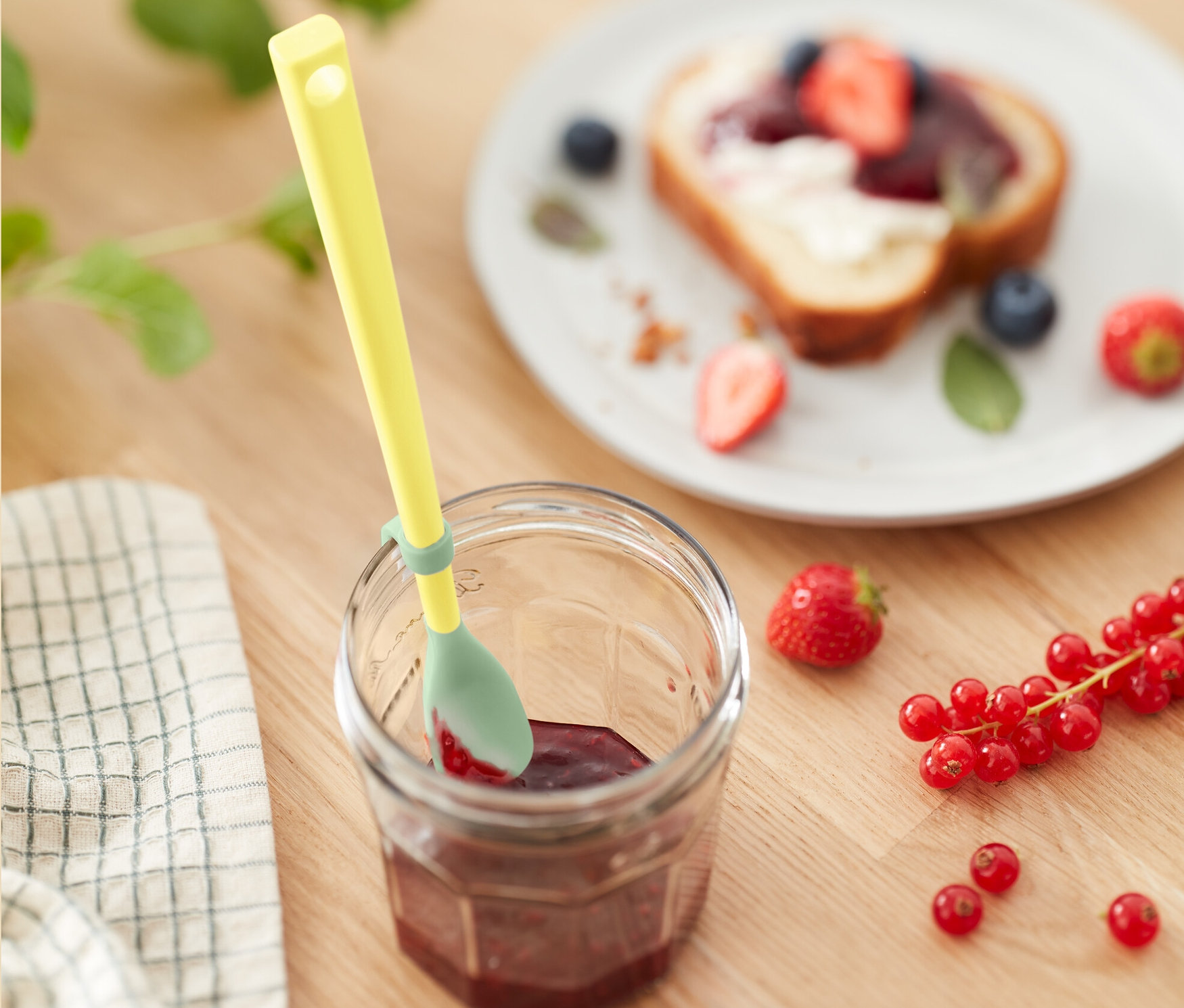 Žlutá silikonová lžíce ve sklenici džemu vedle talíře s toastem s ovocem.