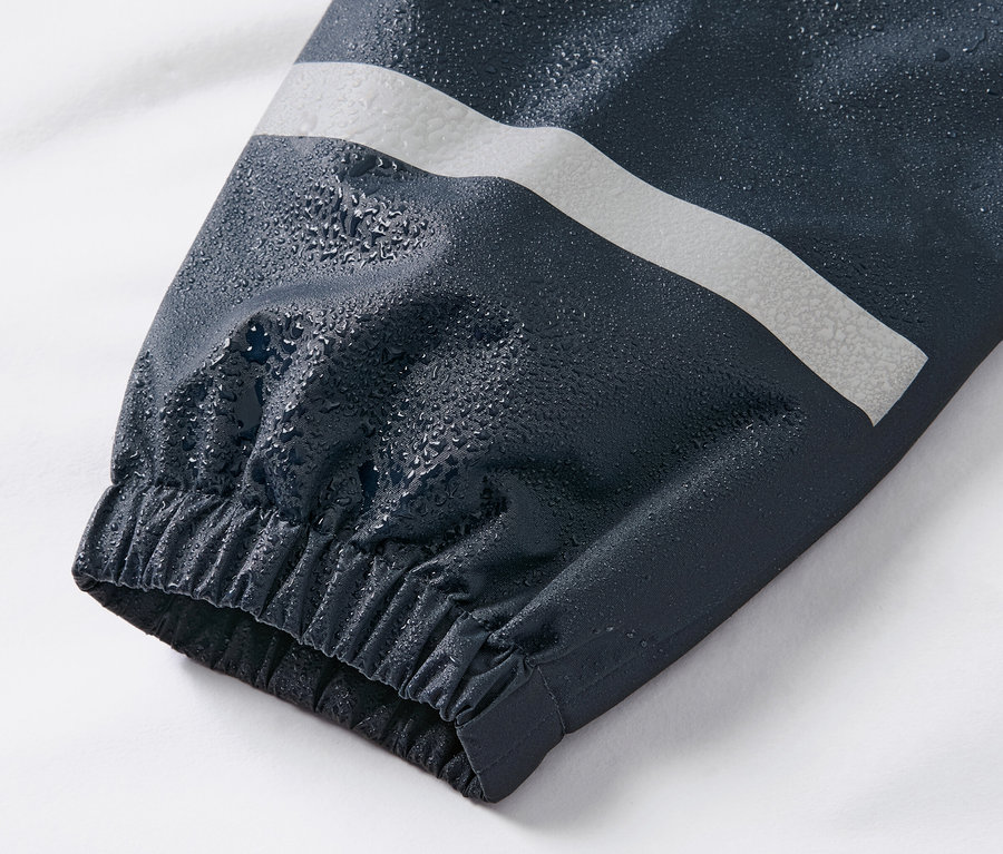 Detailní záběr na rukáv dětských kalhot do deště tmavě modré barvy s reflexním pruhem a kapkami vody.