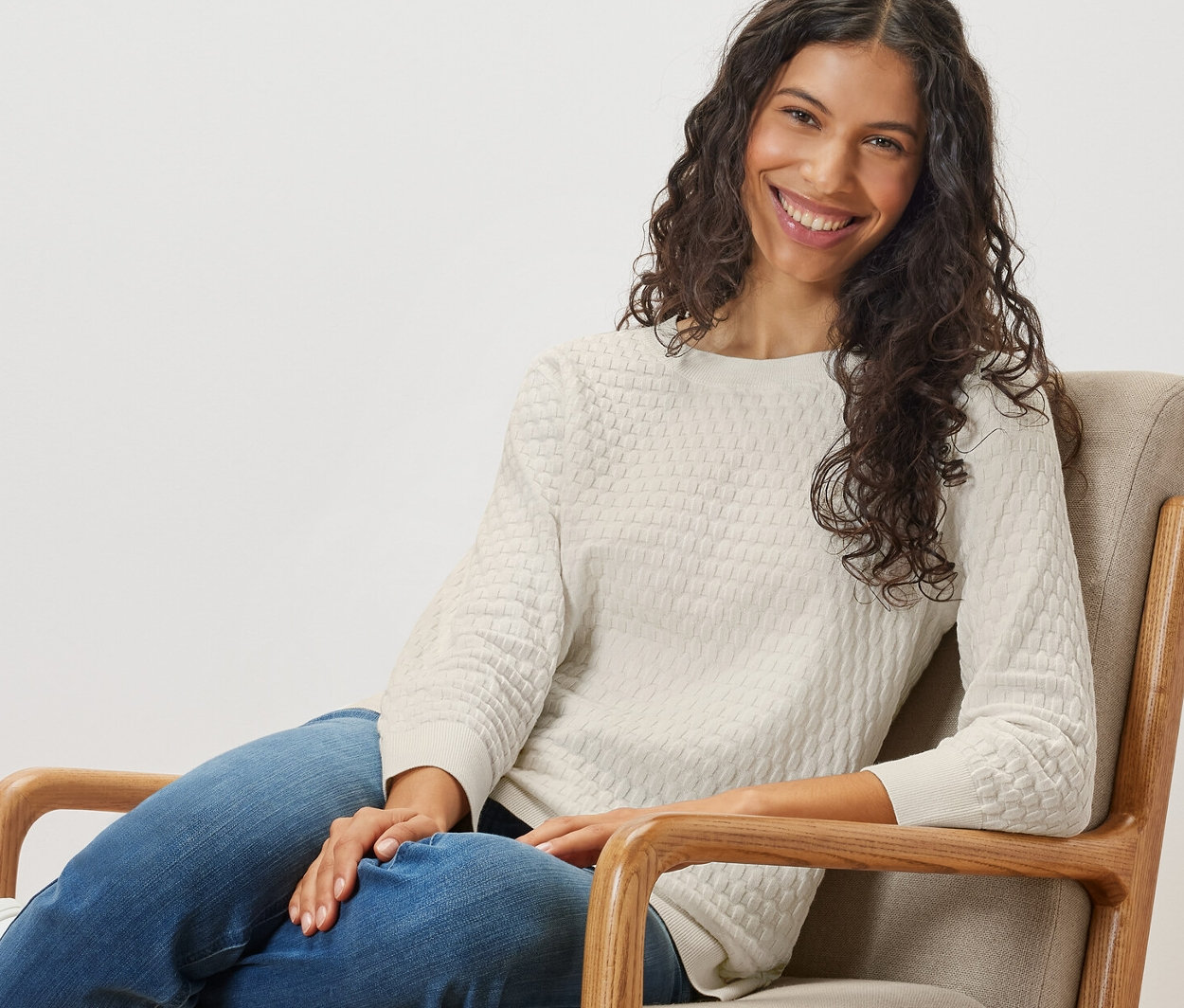 Žena sedí v křesle a má na sobě strukturovaný pletený svetr.