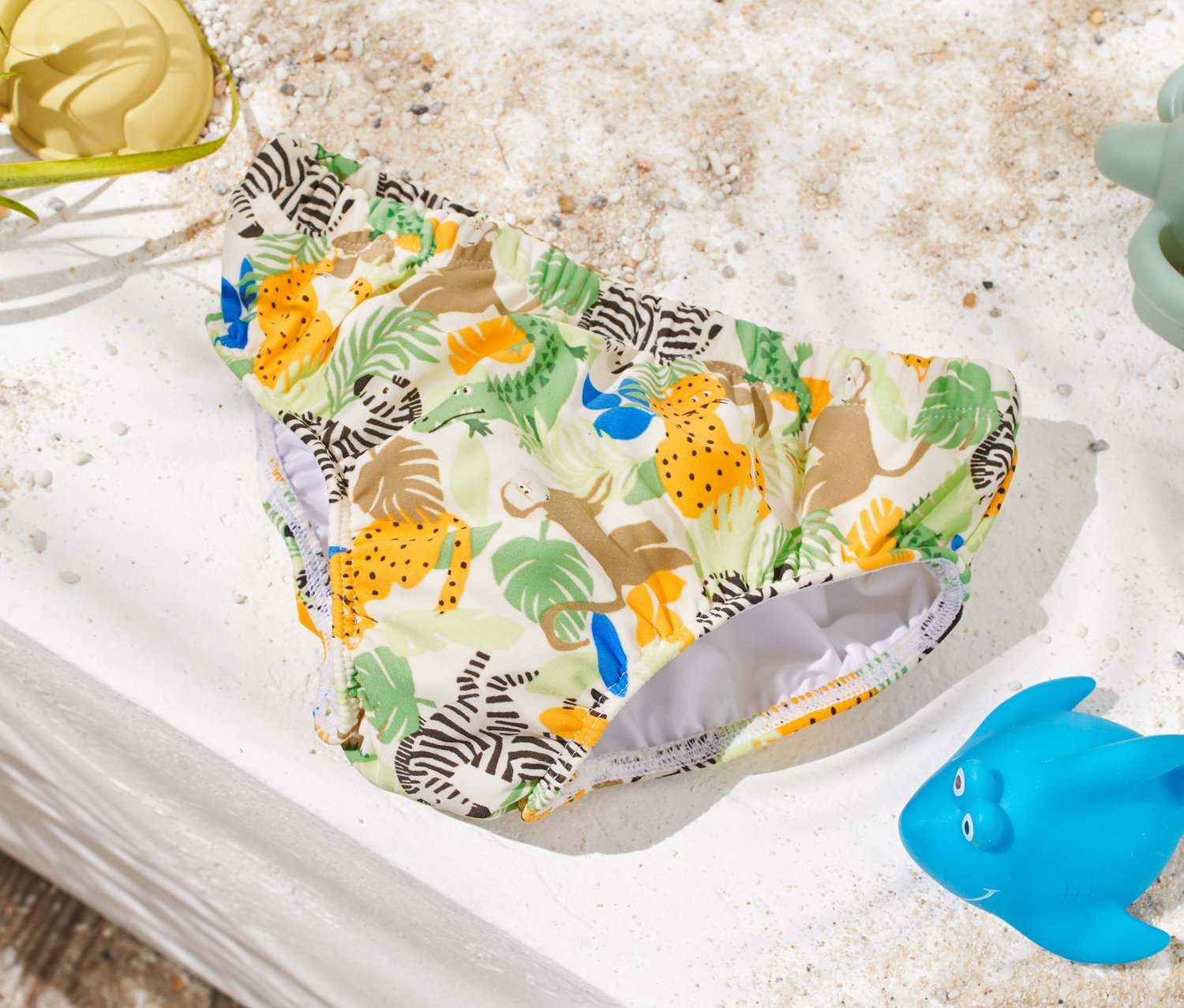 Dětské plenkové plavky se zvířecím potiskem leží na bílém povrchu vedle hraček do písku.