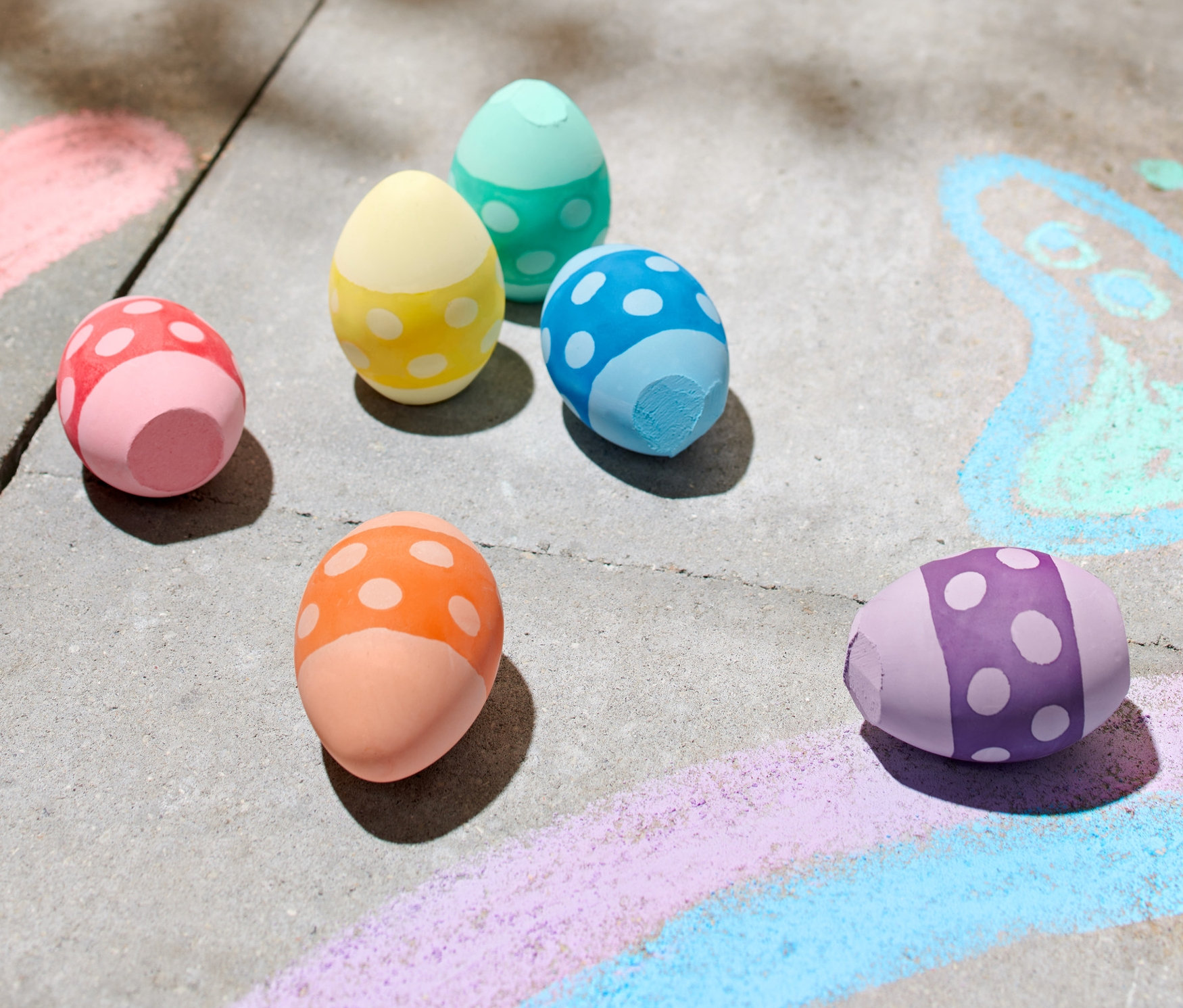 Šest barevných kříd ve tvaru vejce leží na chodníku vedle kreseb křídou.